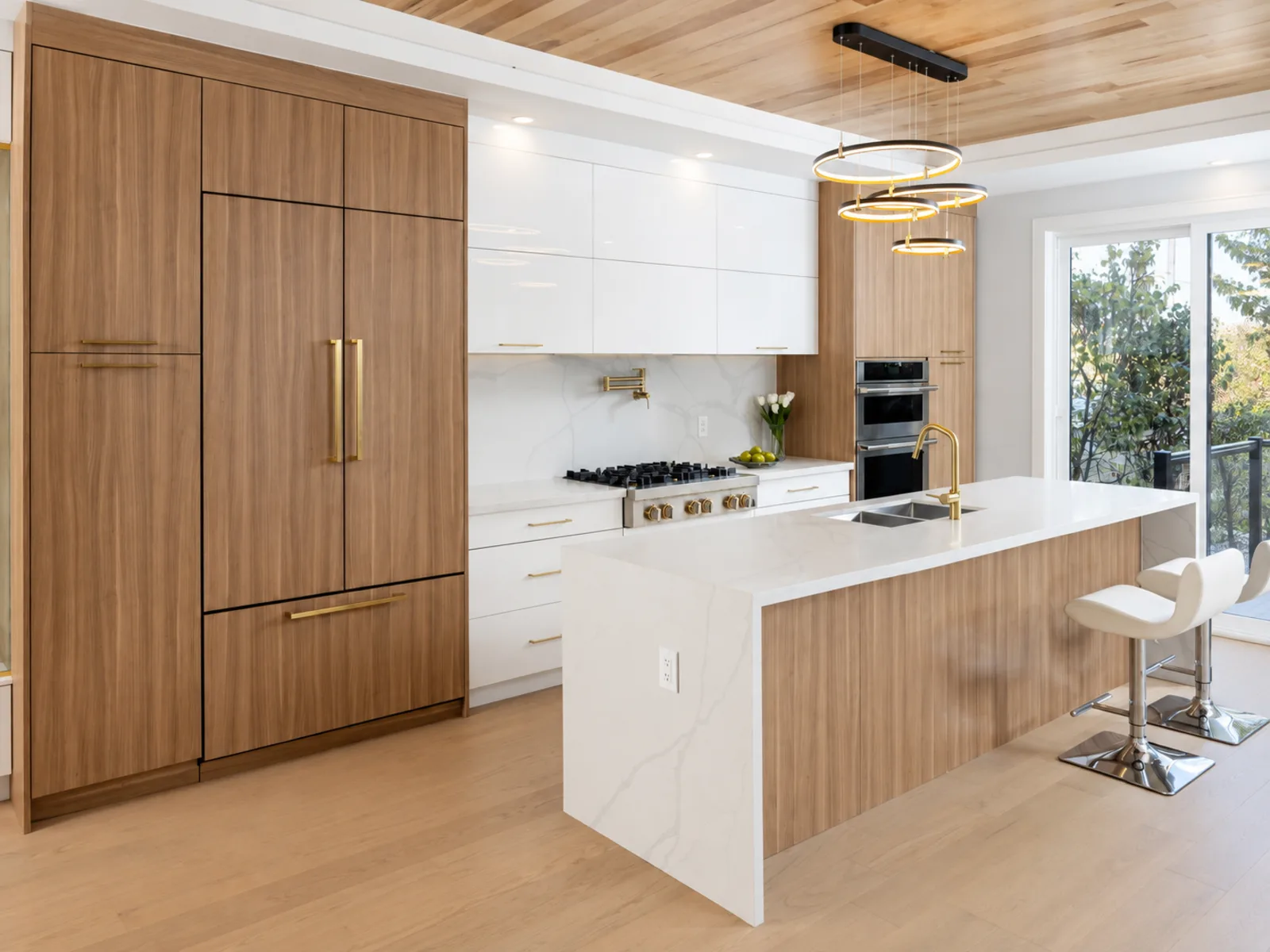 Completed modern kitchen with woodgrain tall cabinets, glossy white uppers, brass-toned pulls and a waterfall island